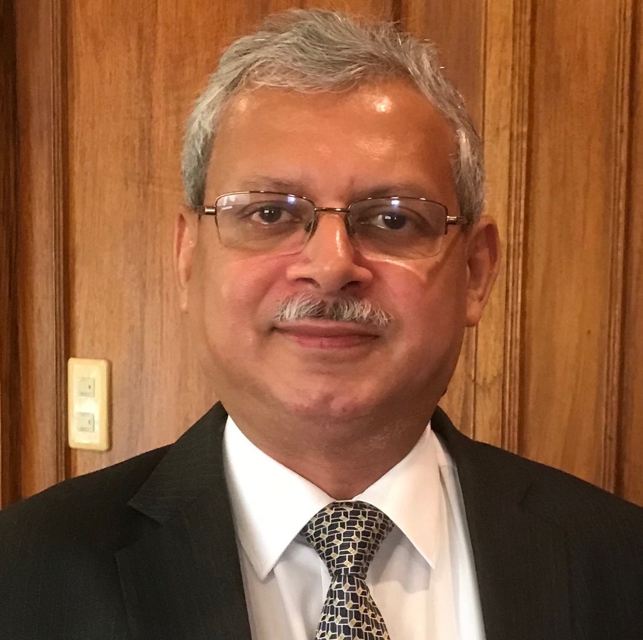 Mr Mazumdr headshot in a dark suit and tie, in front of a wooden background