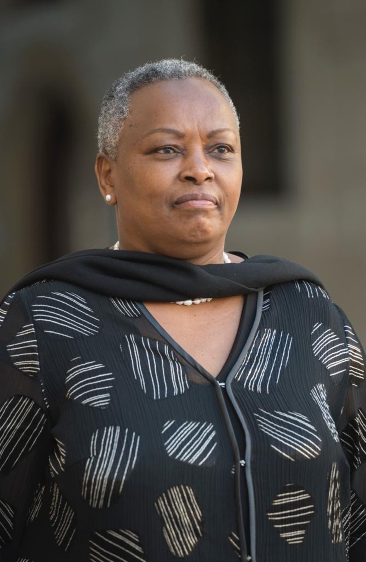 Ambassador Mary Mugwanja is pictured wearing a black dress with white whine diagonal lines and a black shawl. She is smiling and sporting short cropped hair. 