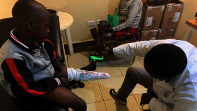 The photo depicts a clinical setting where a healthcare professional is attending to a young boy seated on a chair. The boy appears to have a prosthetic leg, and the professional is seemingly making adjustments or examining the prosthetic device. In the background, another individual is seated, possibly waiting for assistance or accompanying the boy. The image conveys a sense of care and medical support, highlighting themes of assistance and health services accessibility, which are crucial for societal equality and justice. The environment suggests that this may be taking place in an African context based on the appearance of the individuals and the website watermark.