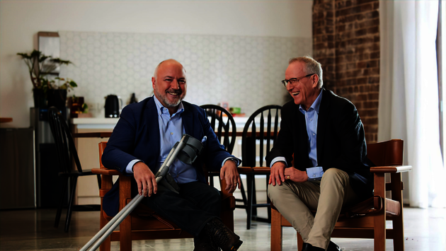 The image shows two men seated next to each other, both smiling and engaged in what appears to be a pleasant conversation. The man on the left has a prosthetic leg, indicating he is a person with a disability. They are dressed in smart casual attire. The setting is indoors with a kitchen area in the background. This photo conveys a sense of inclusion and normalcy in interactions between individuals, regardless of physical differences, promoting themes of equality and tolerance.