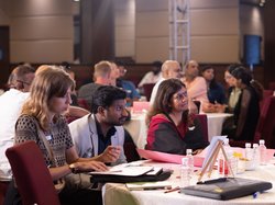 A photo showing the audience seated around large round tables; they appear to be of different nationalities and ethnic backgrounds. At the front, there are three participants taking notes and looking attentively towards the stage; indicating the diverse audience of local and international guests with and without disabilities, and how they attend to learn and exchange ideas and solutions.