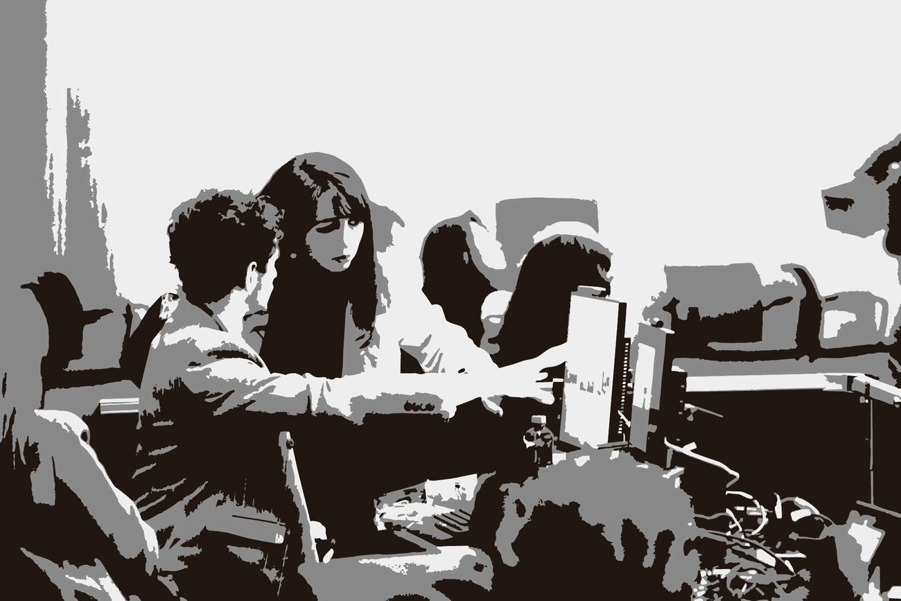 Stylized black, white, and grey image of several people seated around a table during a group discussion, with laptops and other equipment in front of them.
