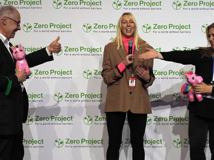 Three people standing in front of a Zero Project branded wall, holding pink stuffed unicorns