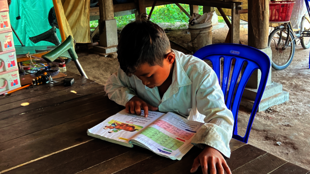 The image shows a young child, likely of Southeast Asian descent, deeply engrossed in reading a book. The child is sitting at a wooden table, leaning forward with attention focused on the page. The environment appears to be a modest, open-air setting, possibly a home or community space, with personal items and a bicycle in the background. The scene evokes a sense of dedication to learning and the importance of education, regardless of one's surroundings. The child's engagement with the book is a powerful reminder of the universal value of literacy and knowledge.