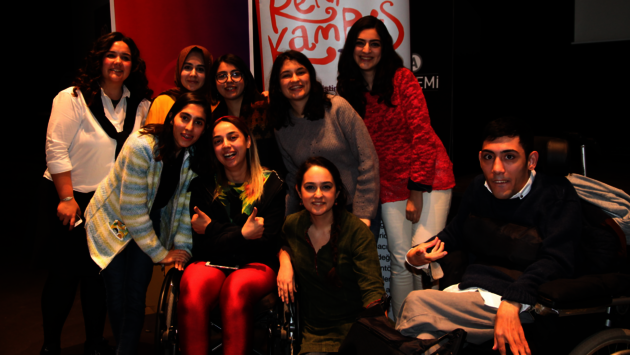 The image shows a joyful group of nine individuals posing together for a photo. Two individuals are using wheelchairs, which signifies the inclusion of people with disabilities. The group is diverse in terms of gender and possibly cultural backgrounds, reflecting a spirit of equality and tolerance. Behind them, a banner with vibrant colors and the words "Renkli Kampus" suggests a context of community, possibly related to an educational or social initiative that promotes diversity and inclusion. The smiles and positive body language of the individuals convey a message of camaraderie and support for one another.