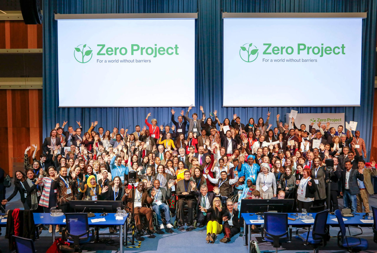 A large group of people laughing and posing in front of a screen with the Zero Project logo at a conference venu