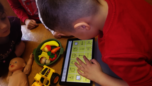 This image shows a group of young children engaged in play and learning activities. A child in a red shirt is interacting with a tablet, possibly using an educational app, while other children are around with toys like a doll and a yellow toy truck. The presence of various toys suggests a playful and inclusive environment that encourages learning through play, irrespective of gender or ability. The children appear focused and are likely developing important cognitive and social skills in a supportive setting.