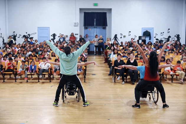 This image shows a diverse group of schoolchildren watching a performance in a school auditorium. In the foreground, two persons in a wheelchair appears to be engaging in an expressive dance or performance art, whilst they are flanked by two standing dancers who are equally dynamic in the expressions with their hands extended into the air in a V-like shape. The audience looks captivated and surprised by the performance. The scene promotes inclusivity and the idea that art and expression are accessible to all, regardless of physical abilities. The performers demonstrate courage and creativity, which serves as an inspiration to the audience, highlighting themes of equality and the celebration of diverse abilities.