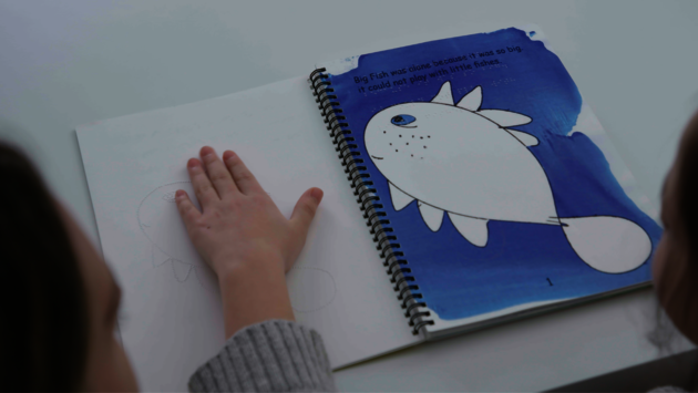This image shows a close-up of an open book with a tactile picture that a child is exploring with their hand. The page visible contains a large printed fish and text that reads "Big Fish was alone because it was so big. It could not play with little fishes." The tactile image is a raised outline of the same fish, allowing a child, possibly with visual impairments, to feel and understand the picture through touch. The book appears to be an educational tool designed for inclusive learning, promoting accessibility and catering to the needs of children with different abilities. The image embodies themes of understanding, inclusivity, and the importance of providing equal opportunities for learning and engagement for all children.