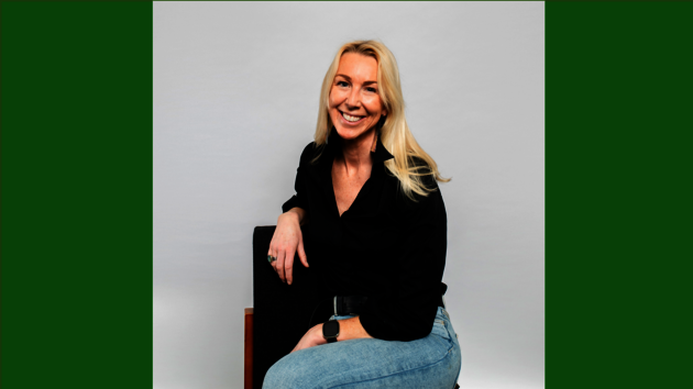 A woman with blond hair, a black button down shirt, and jeans, sitting legs crossed in a chair and smiling.