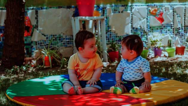 The image captures a tender moment between two young children sitting on a colorful play mat outdoors. The mat features vibrant sections of red, green, blue, and yellow, and the children are engaging with each other, suggesting a sense of curiosity and friendship. The background is a garden setting with a variety of plants and decorative elements, creating a peaceful and playful environment. The children's expressions and body language reflect innocence and the universal nature of childhood, transcending any specific cultural or geographic origin. The scene promotes themes of equality and tolerance, as the children interact without prejudice, exemplifying the pure and accepting nature of early childhood interactions.
