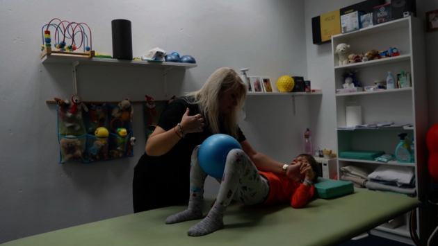 This image depicts a therapy session where a healthcare professional, a woman, is assisting a young child. The child is lying on a therapy mat with a blue exercise ball placed between their legs, which the therapist is holding. The environment suggests a pediatric therapy clinic, evidenced by the toys, therapy equipment, and child-friendly decorations around the room. The therapist is focused on the child, indicative of a caring and supportive interaction aimed at aiding the child's development or rehabilitation. The scene embodies themes of assistance, care, and the nurturing of growth and improvement, all within a setting designed to be welcoming and engaging for children.