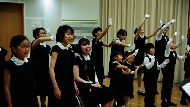 The photo depicts a diverse group of Asian children and a young adult engaged in a choral performance. They are dressed uniformly in black attire with white gloves, enhancing the visual harmony of the group. A young woman in a wheelchair is prominently included among the standing children, illustrating inclusivity and the normalization of disability within the community. The expressions on their faces range from concentration to joy, suggesting a shared experience of music and performance that transcends individual differences. The setting appears to be a well-lit, indoor space, likely a school or community center, fostering an environment of learning and cultural expression.