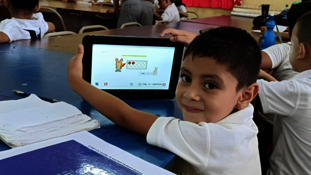This image shows a young child in a classroom environment, proudly holding up a tablet displaying an educational activity. The child is smiling and looking at the camera, conveying a sense of joy and engagement with the learning process. Around the child are other students, focused on their work, and school materials such as notebooks are visible on the tables. This scene highlights the importance of access to technology in education and the positive impact it can have on a child's learning experience. The atmosphere suggests a collaborative and inclusive educational setting where every student has the opportunity to learn and grow.