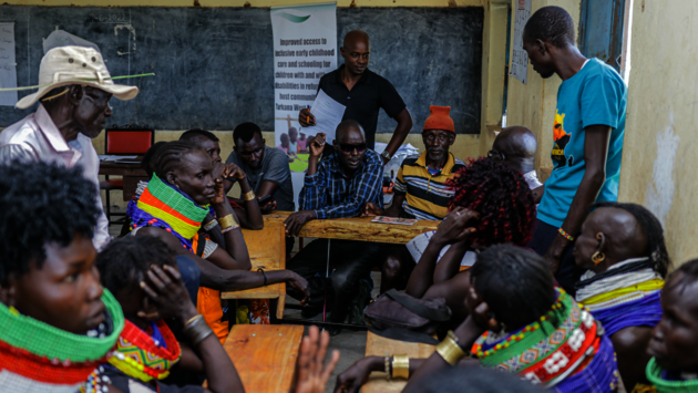 The photo depicts a group of African men and women gathered around tables in a community setting, possibly a classroom or meeting space. The individuals are engaged in what appears to be a discussion or workshop, with a focus on inclusivity and community support, as suggested by the poster in the background mentioning improved access to inclusive early childhood care. The attire and adornments of the participants, along with the context provided by the poster, suggest a commitment to cultural identity and social development. The scene embodies themes of collaboration, education, and empowerment within a diverse community.