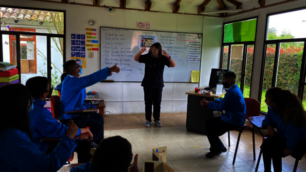 This is a classroom setting with a diverse group of students wearing blue school uniforms, attentively participating in a lesson. A teacher stands at the front, gesturing as part of the instruction, demonstrating an active learning environment. The whiteboard behind the teacher is filled with written educational material, suggesting the subject might involve language learning. The room is well-lit with natural light, creating a positive and engaging atmosphere. The students are engaged in the learning process, with one student pointing towards the board, indicating participation and interaction. The scene reflects an inclusive educational environment where learning and student engagement are prioritized.