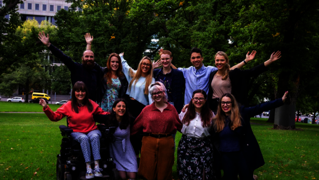 This is a joyful and diverse group of individuals posing together in a park. They are smiling and several are raising their arms in celebration or greeting. The group is casually dressed, suggesting a relaxed and friendly atmosphere. One person is using a motorized wheelchair, illustrating inclusivity and the representation of people with disabilities. The lush greenery in the background adds to the sense of community and connection to nature. The image conveys themes of happiness, diversity, inclusion, and equality, as the group appears united and supportive of one another.
