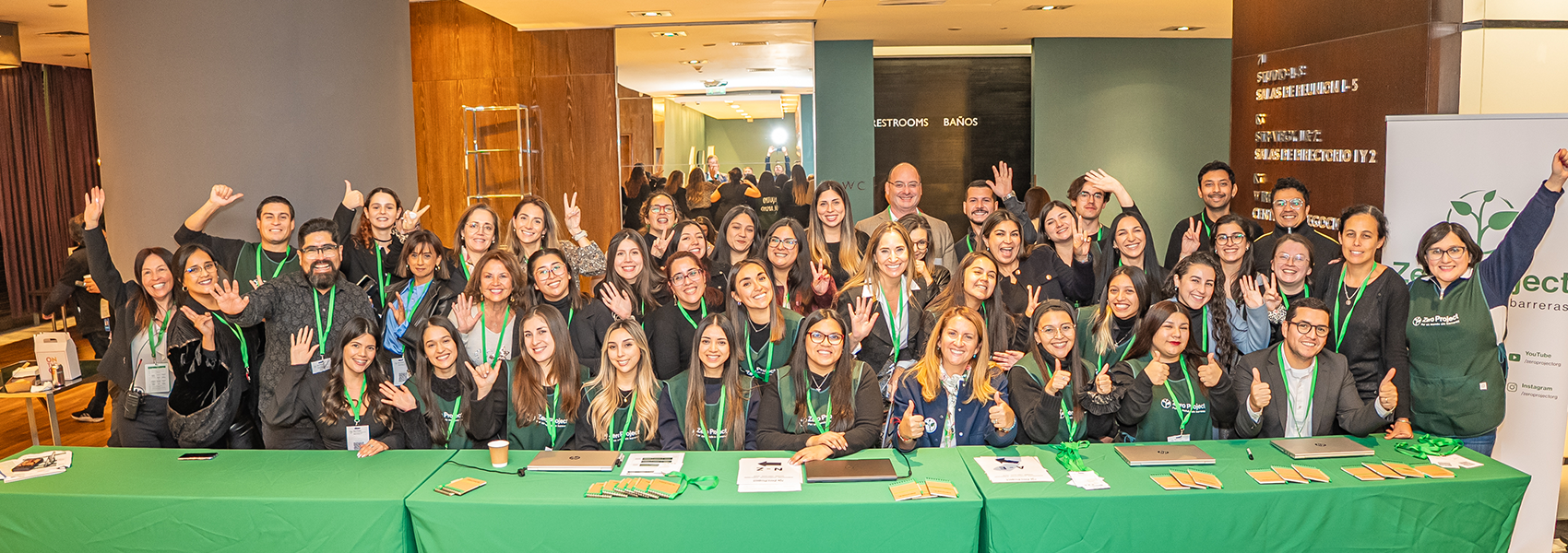 A large group of people from the organizing team of the Zero Project 2025 conference pose smiling behind a green table with the initiative's logo. All are wearing name badges and green lanyards, in a hotel or conference center setting.