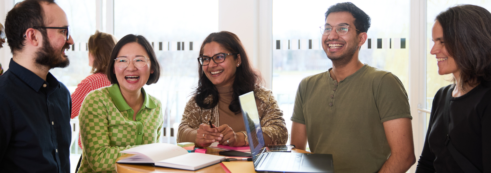 A diverse group of five people in a casual business setting around a high table with a laptop; laughing and smiling.