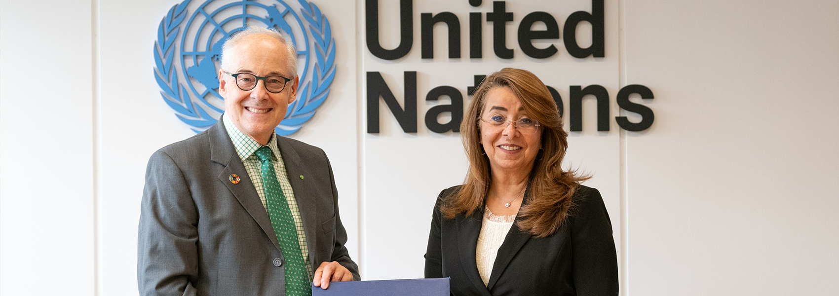 Martin Essl and Ghada Waly standing and smiling in front of the United Nations logo and sign. They are holding a blue folder with the UN emblem. Martin Essl, on the left, is wearing glasses, a gray suit, and a green tie. Ghada Waly, on the right, is wearing glasses, a black suit with a gold buckle belt, and a white blouse. They are positioned behind a table with two pens on a stand.